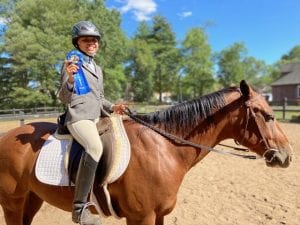 A girl on a bay horse smiling and holding up a blue first place ribbon at a horse show