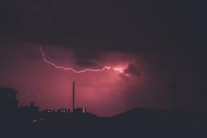 A bolt of pink lighting flashing across and illuminating a dark sky. There are silhouettes of buildings and landscape in the background.