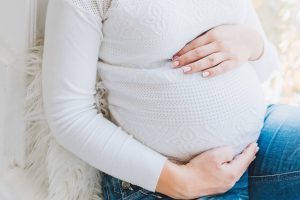 A pregnant woman sitting on a windowsill in a white shirt and jeans.