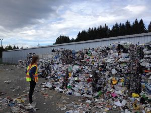 A woman in a yellow high visibility safety vest standing in front of cubes of plastic waste. A blue building and pinetrees are in the background.