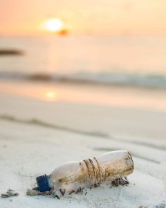 A plastic bottle with a blue cap on a beach with the ocean in the background at sunset.