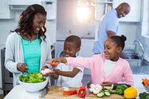 Mother and 2 kids making a salad with the dad it the background smiling