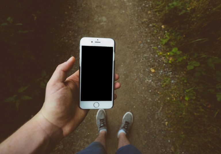 a hand holding a cell phone above bare ground and shoes