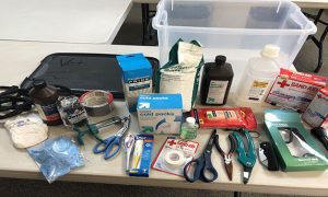 A variety of medical supplies including tools, bags, and bottles on a table in front of an open clear plastic bin.