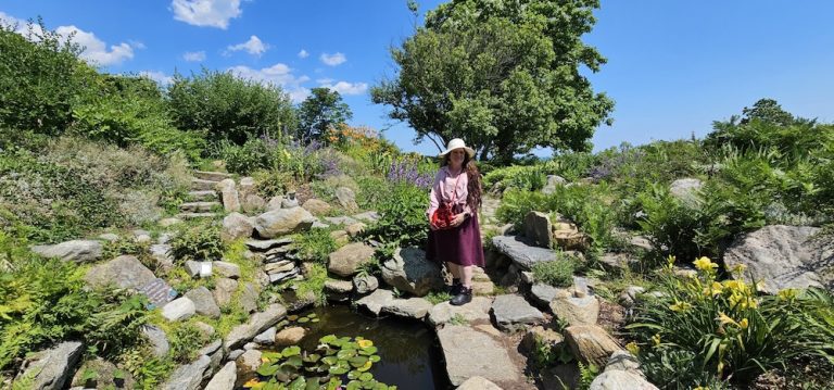 a woman standing in a garden in front of rocks with a large tree behind her and green leaves on it