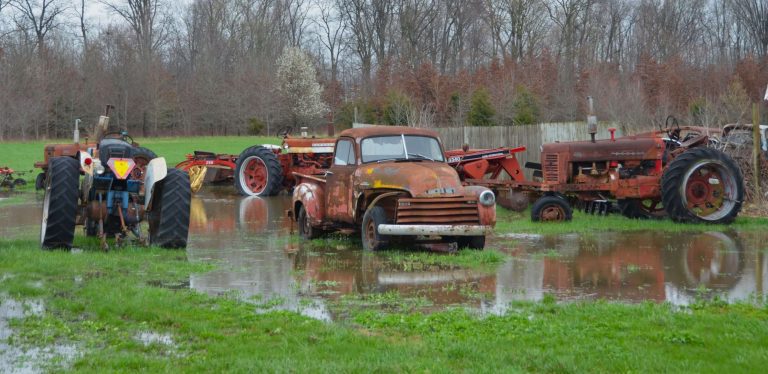 A photo d red rusty tractors and cars sitting in a flooded field with a brown fence and trees in the background.