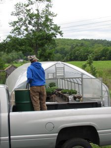 A person in a blue hoodie standing in the bed of a gray pickup truck, holding a black hose into a green metal barel, in front of a greenhouse with plants inside.